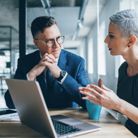 Two senior colleagues, male and female presenting, discuss something in front of a laptop